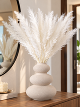 Beige textured ceramic flower vase with white pampas grass displayed on a marble table.